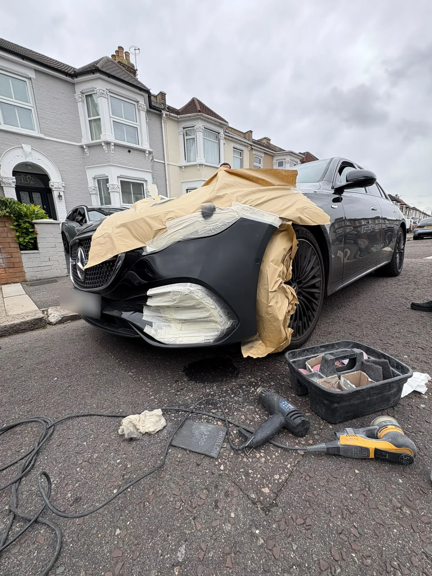 The Mercedes E-Class masked up with paper and tape while the front bumper is prepped and painted on the customer's street